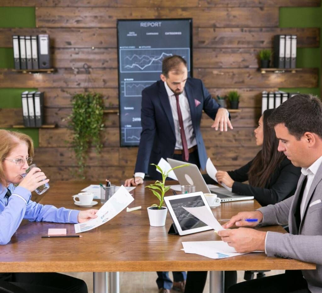 Business meeting in a wood-paneled conference room, with people reviewing documents and charts around a table.
