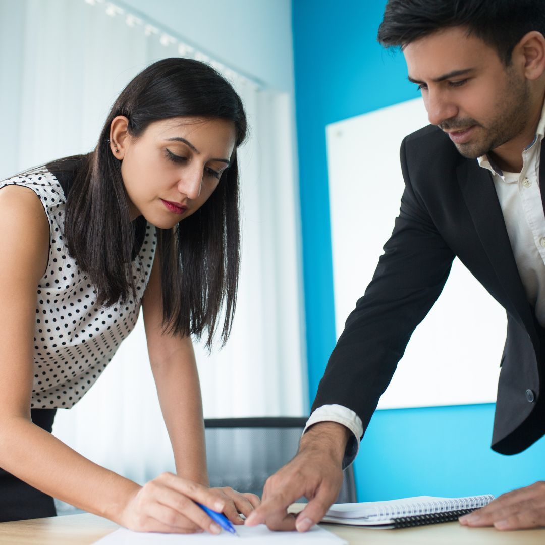 Two coworkers lean over a table, reviewing papers and writing notes in a bright office.