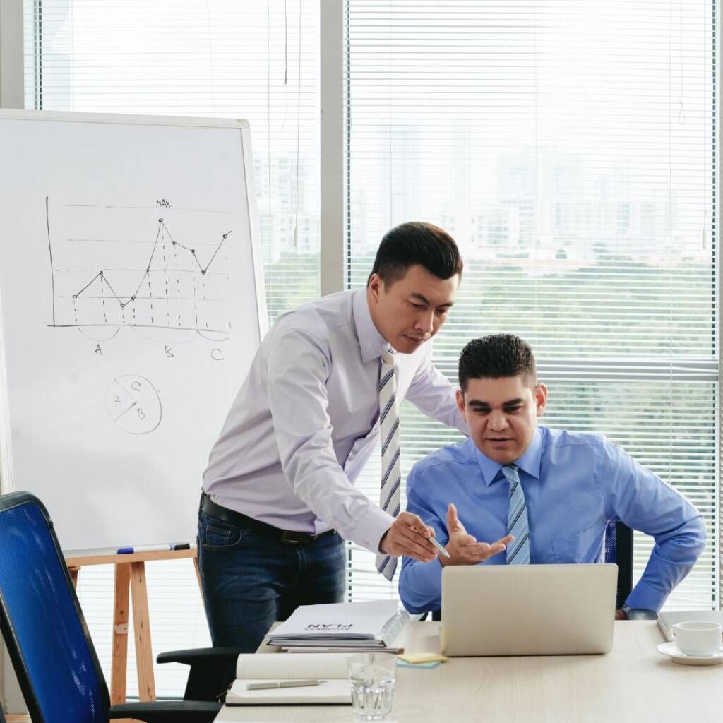 Two coworkers reviewing a laptop in a bright office, with charts on a whiteboard nearby.