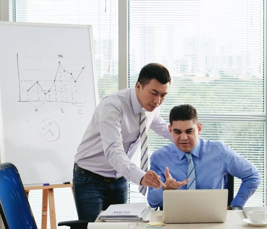 Two coworkers discussing a laptop in a bright office, with a whiteboard chart in the background