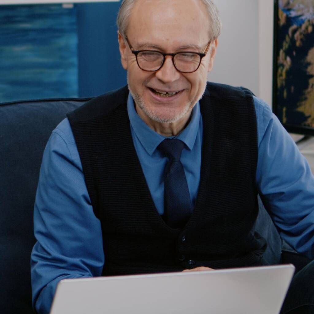 Smiling man in blue shirt and black vest working on a laptop on a couch