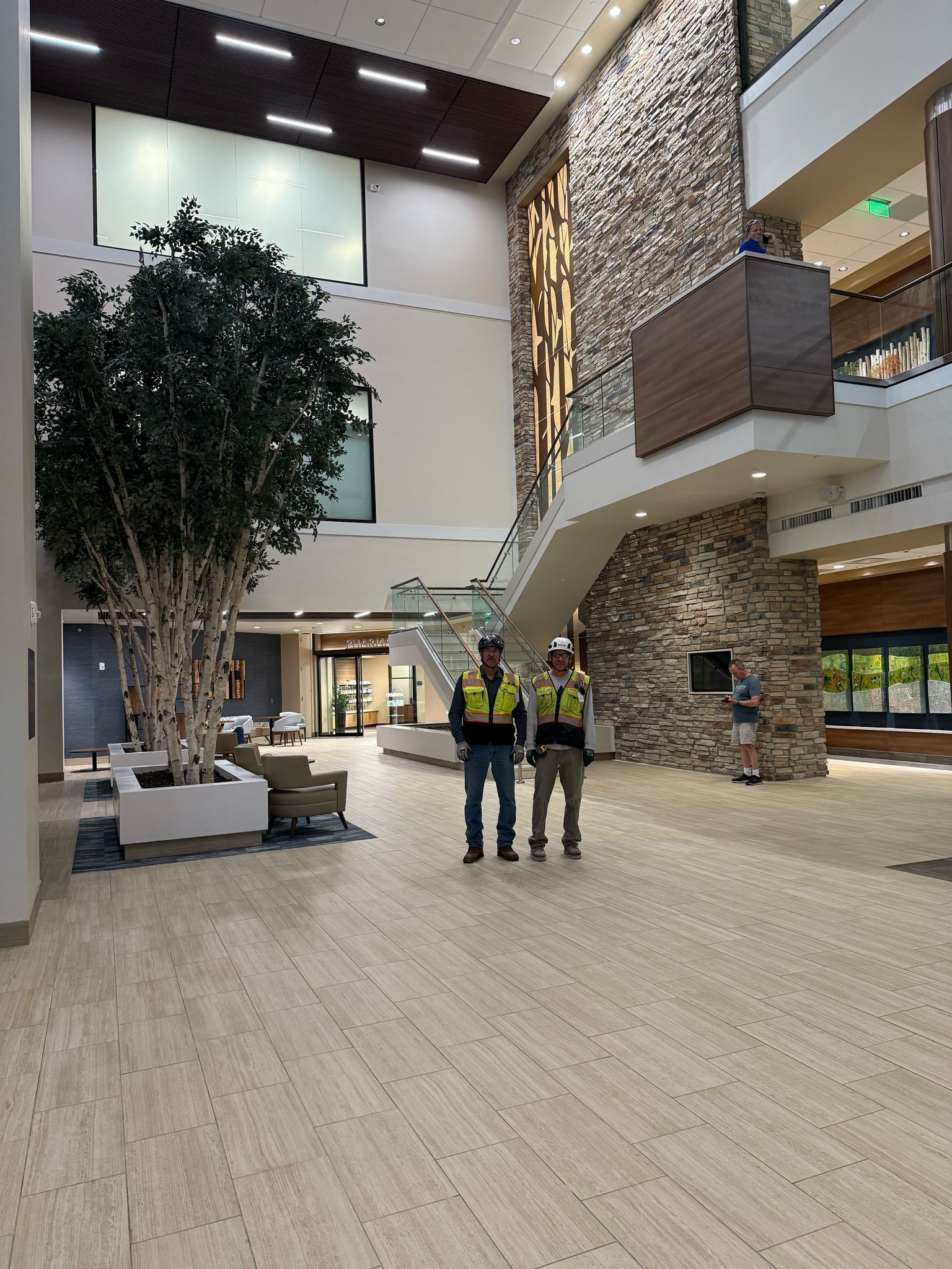 Two people in safety vests stand in a building's lobby with a large tree and stone wall.