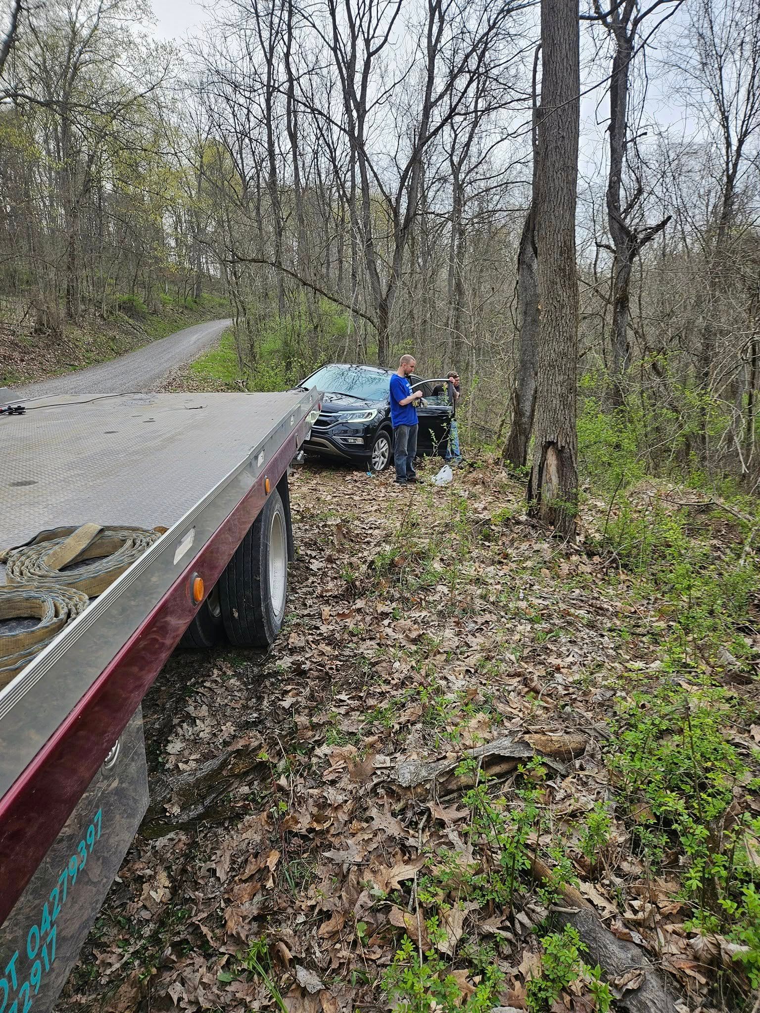 A tow truck loading a black car beside a wooded road; a person stands near the car.
