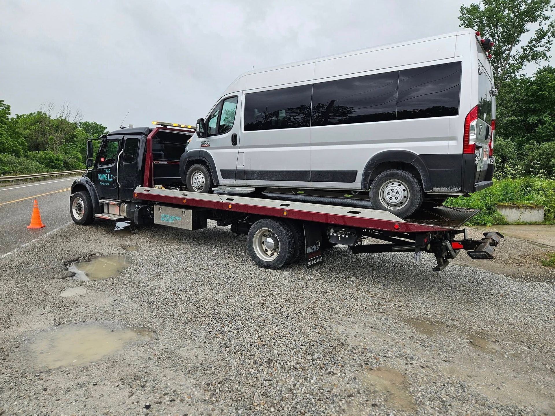 Tow truck carrying a silver passenger van on a roadside with wet gravel, overcast sky.