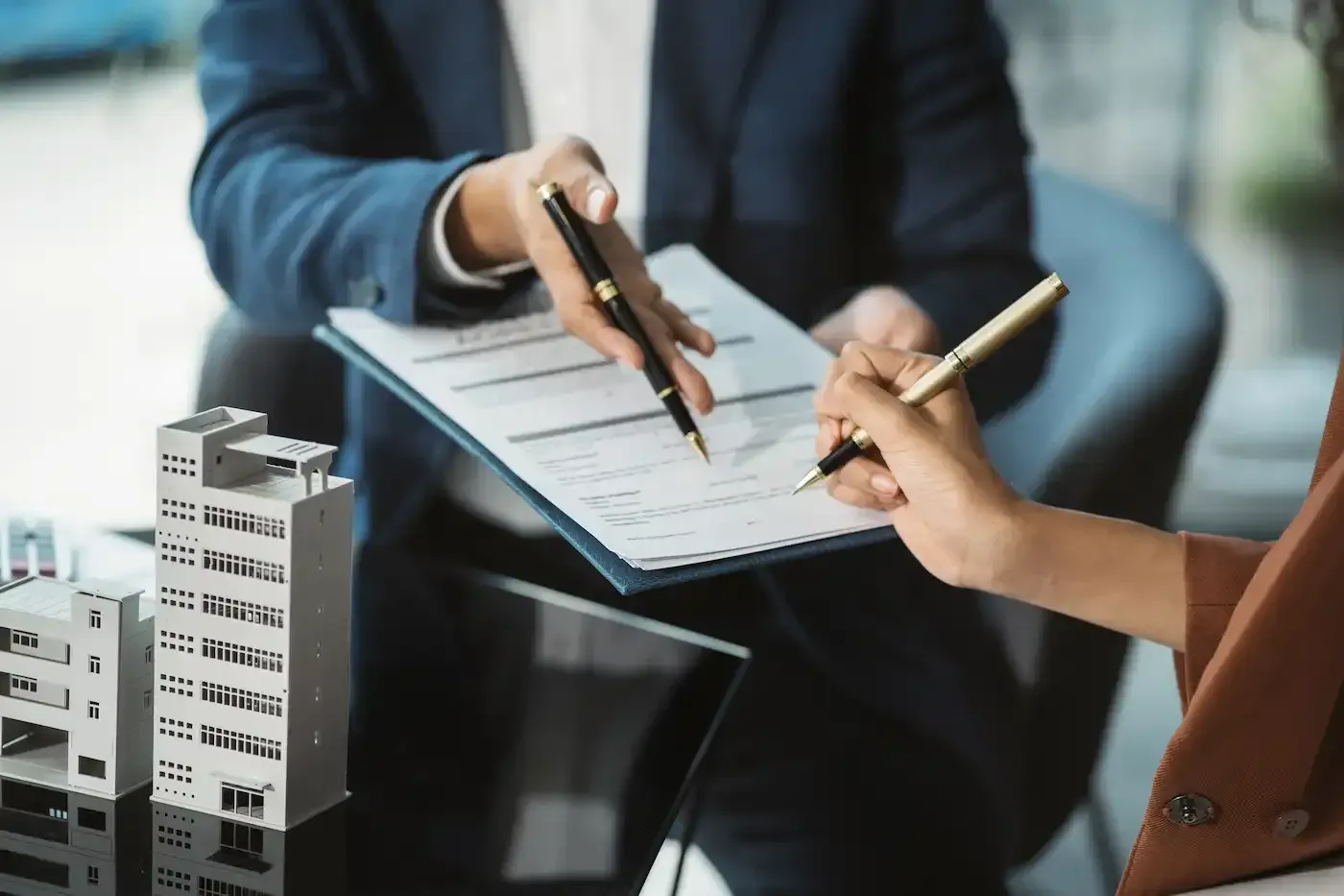 Two people signing a contract on a clipboard, with small building models on the desk.