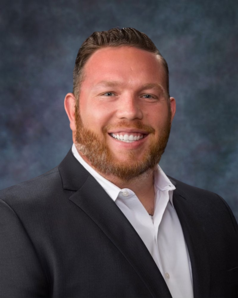 Man in black suit and white shirt, smiling with a beard, against a dark blue background.