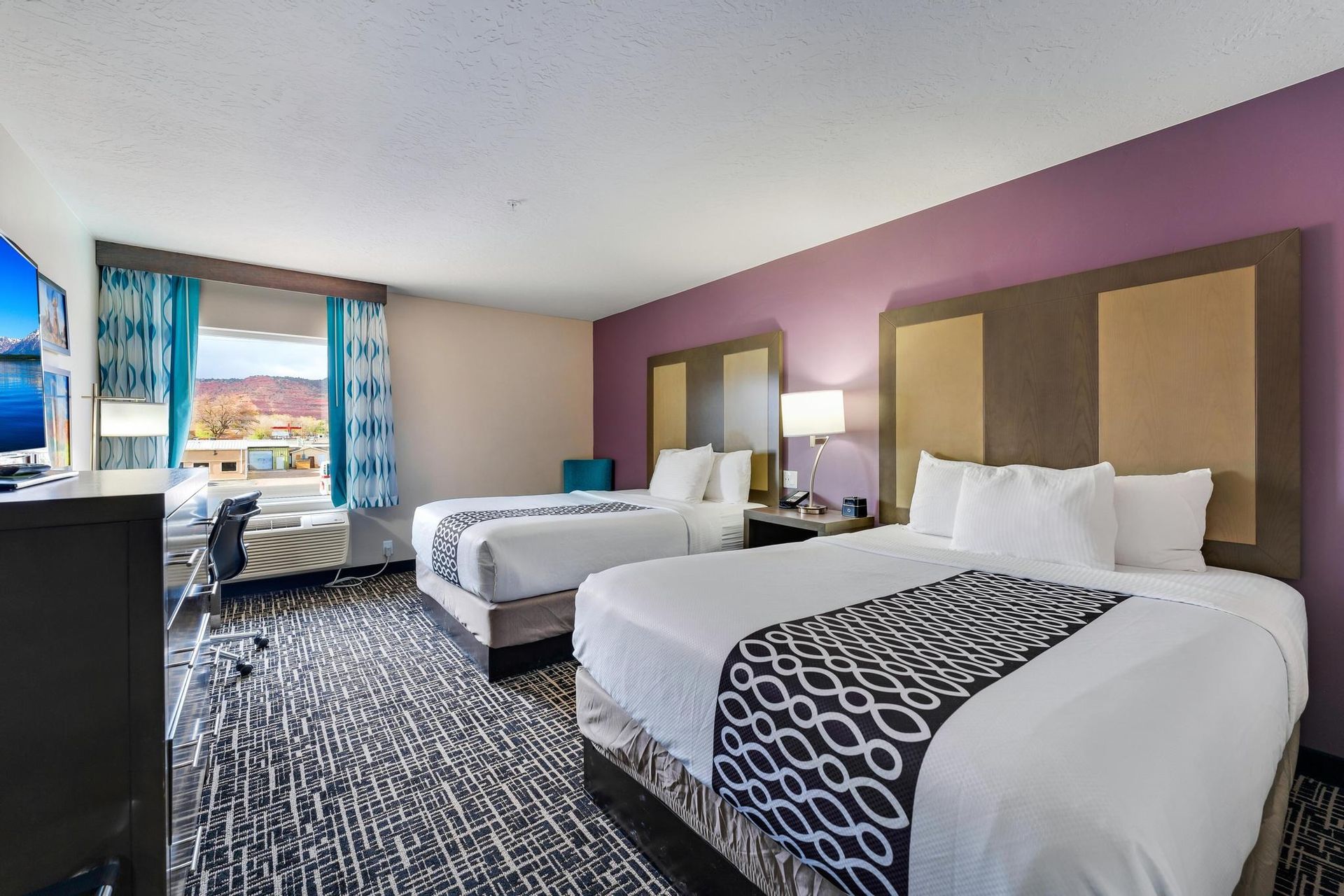 Hotel room with two queen beds, purple wall, view of scenery, desk, and patterned carpet.