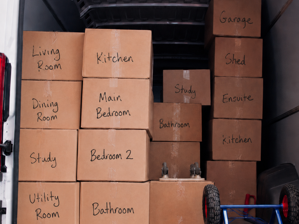 Cardboard boxes stacked on a porch, labeled with room names, beside stairs and a bicycle wheel.