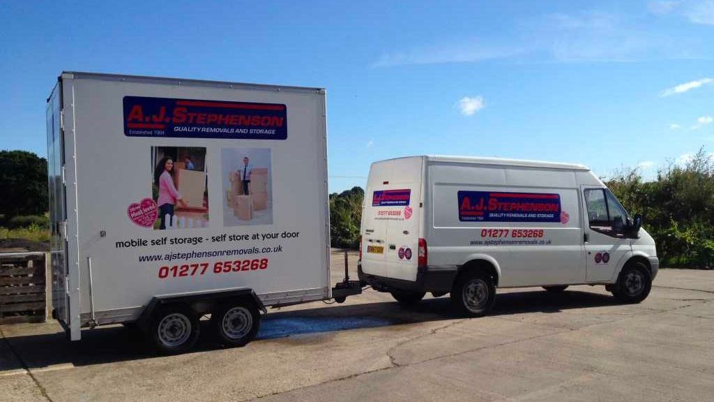 White van towing a trailer with business logos and contact information against a blue sky.