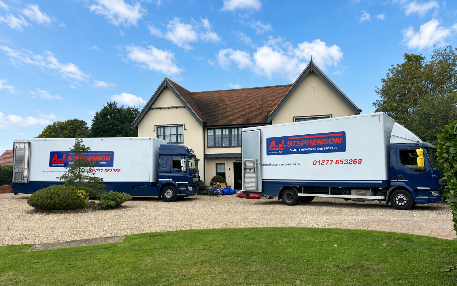 Two white moving trucks parked in front of a house with a red-tiled roof and gravel driveway