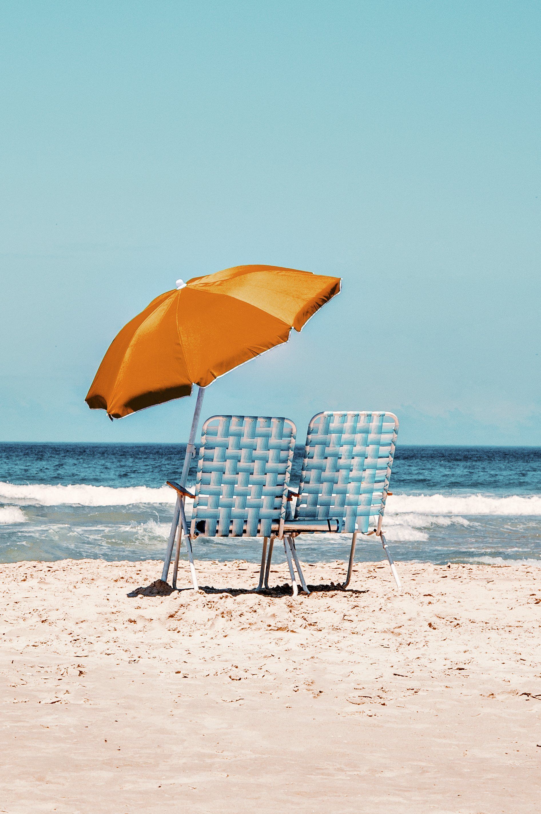 Orange beach umbrella beside two blue striped chairs on a sunny sandy beach with ocean waves.