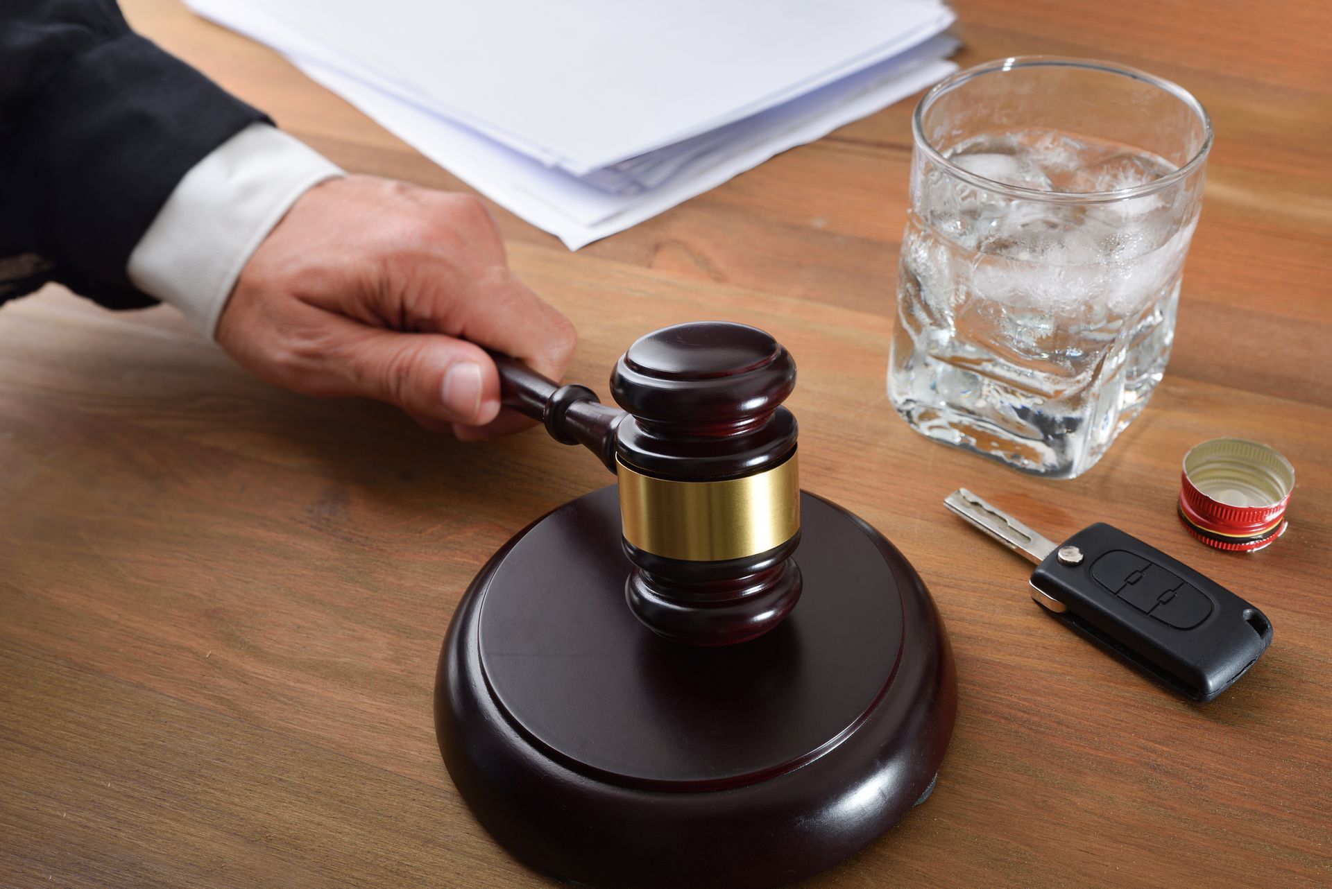 Gavel on a stand, hand in suit jacket, glass of alcohol, car key, and alcohol bottle cap on a wooden table.