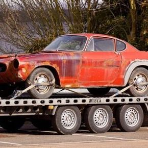 Rusted red vintage coupe strapped on a car trailer with three wheels visible