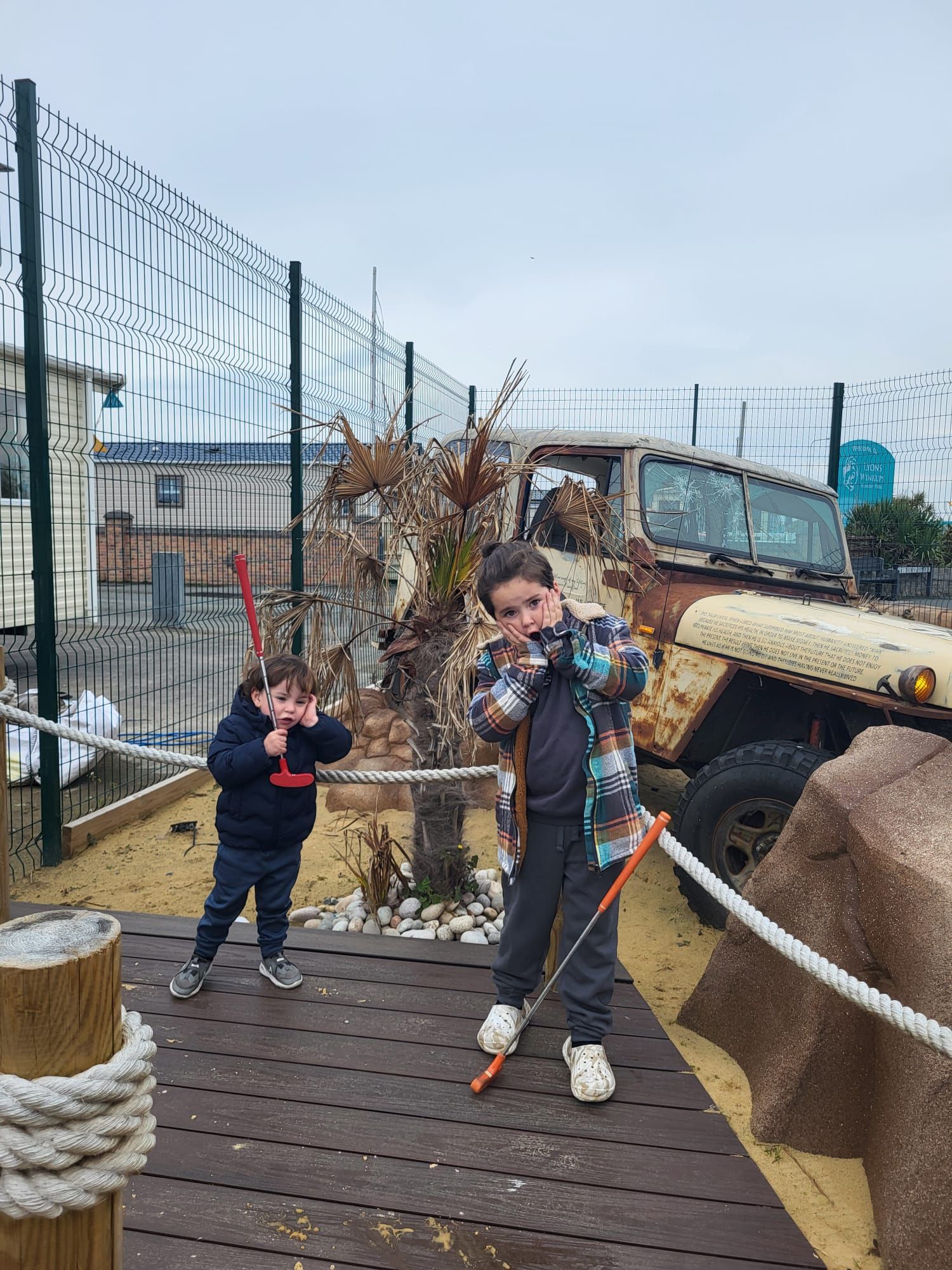 Two children pretending to shoot, standing on a boardwalk near an old vehicle, cloudy day.