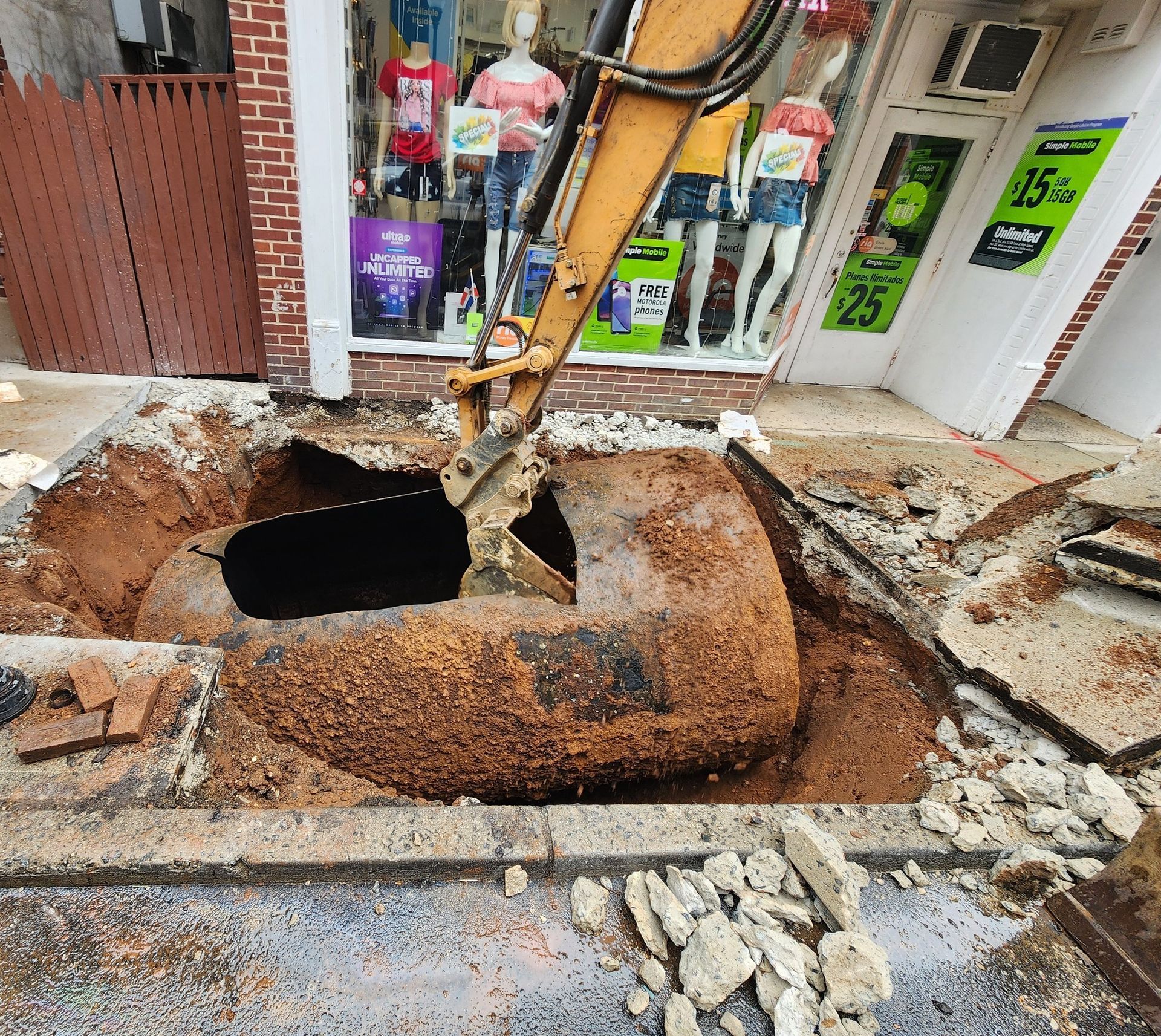 Excavator removing a buried metal tank in front of a store with mannequins in the window. Brown dirt and debris.