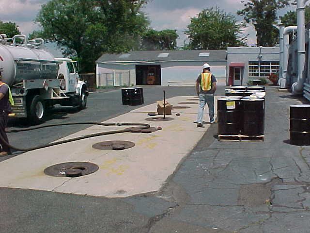 Truck pumping liquid, workers near barrels and manhole covers on concrete pad.