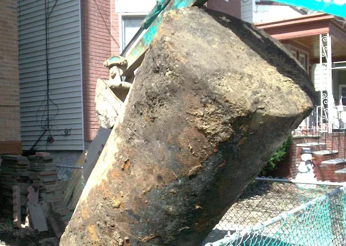 Excavator bucket filled with dark soil next to a building, fence in foreground.
