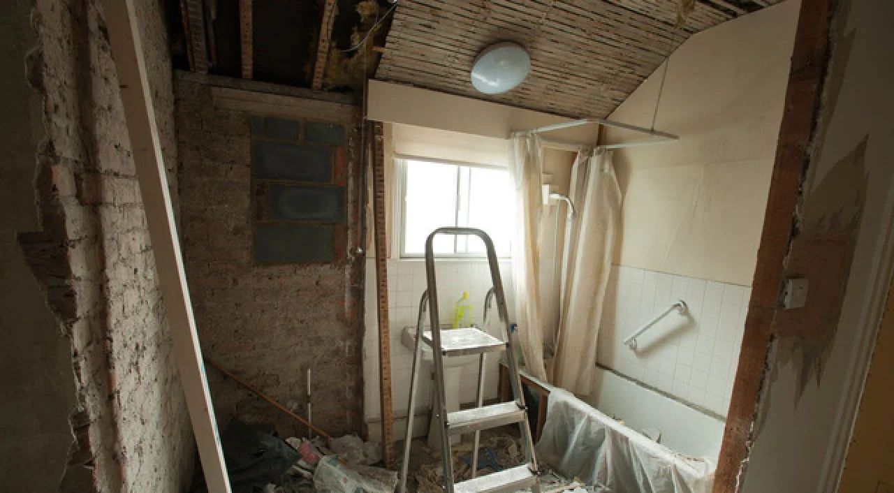 Damaged bathroom under renovation with exposed brick, debris, ladder, and shower curtain.