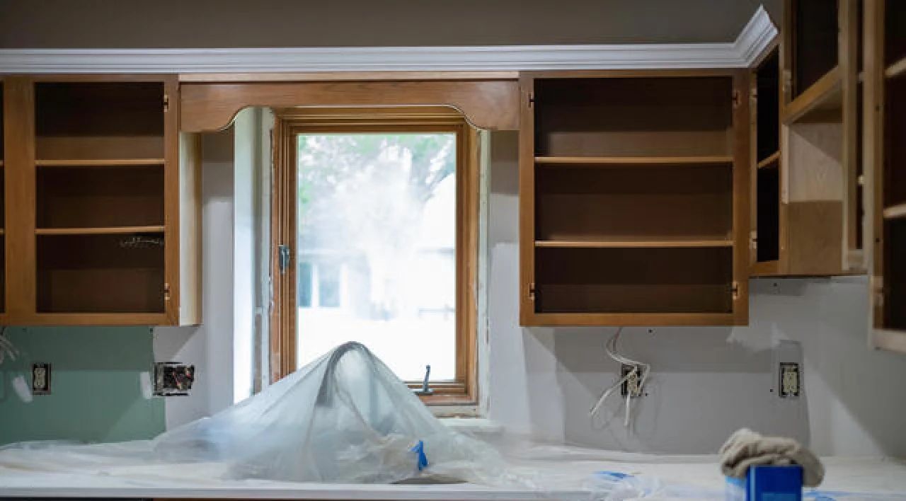 Kitchen under renovation with open wooden cabinets, a window, and a counter covered in plastic sheeting.