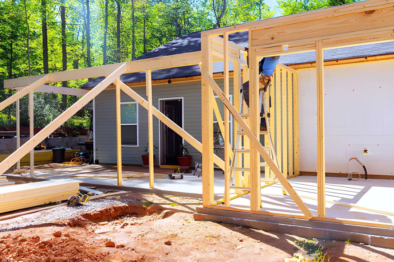 Wood-framed house under construction with roof trusses and plywood siding in a sunny wooded area