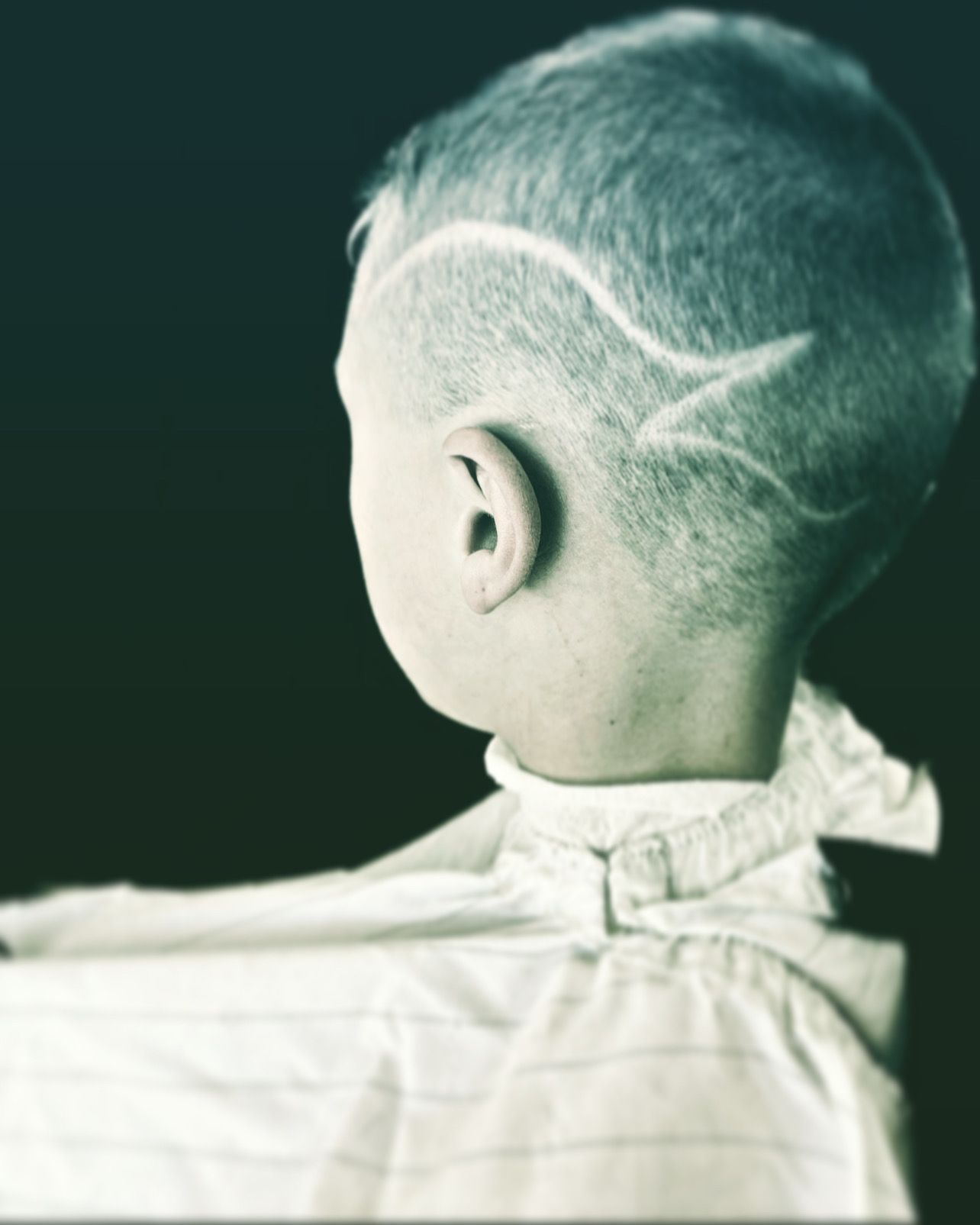 Boy with a shaved haircut featuring a curved design on the side of his head. He's wearing a white shirt.