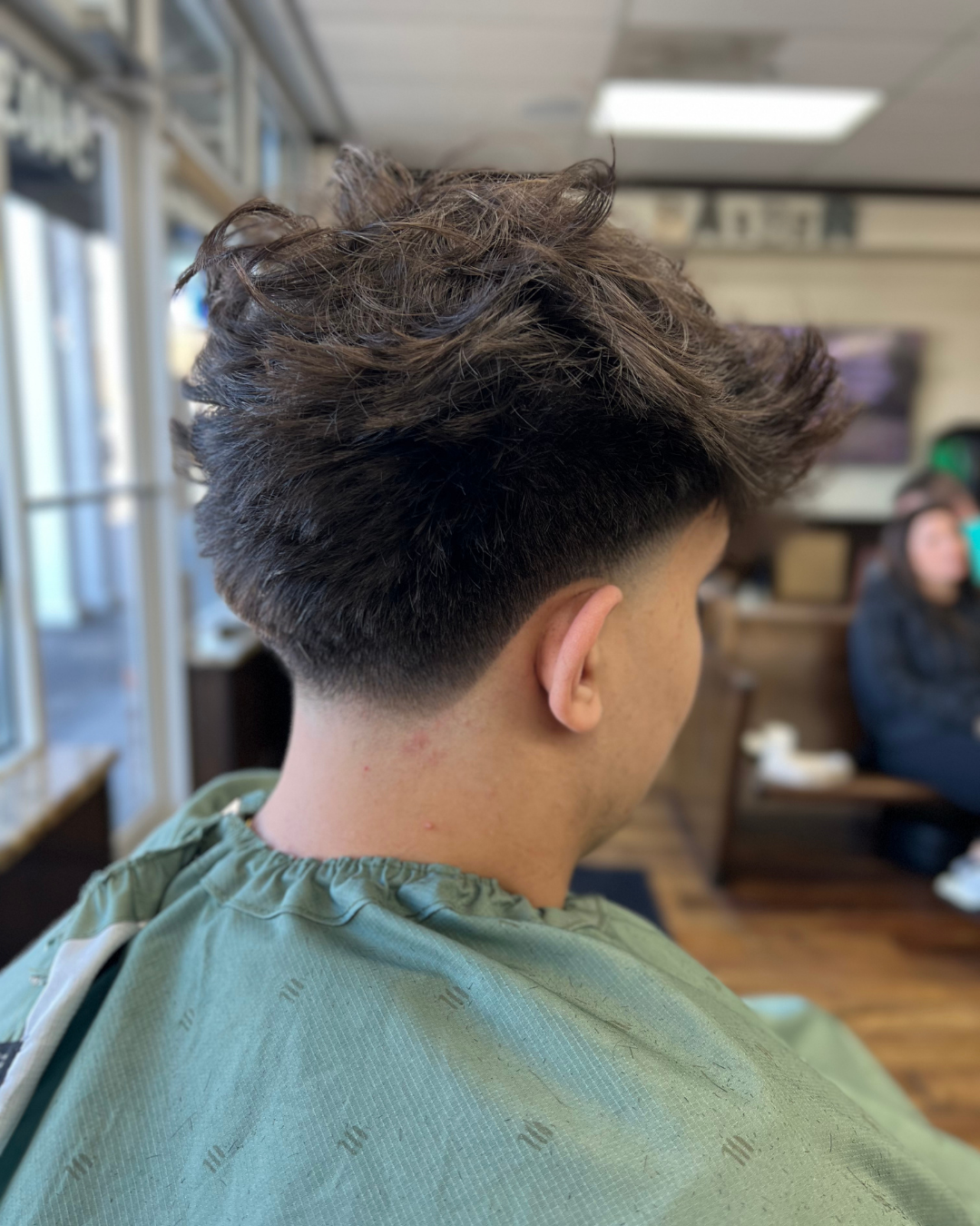 Man with textured, dark hair, wearing a barber's cape, fresh haircut with a fade.