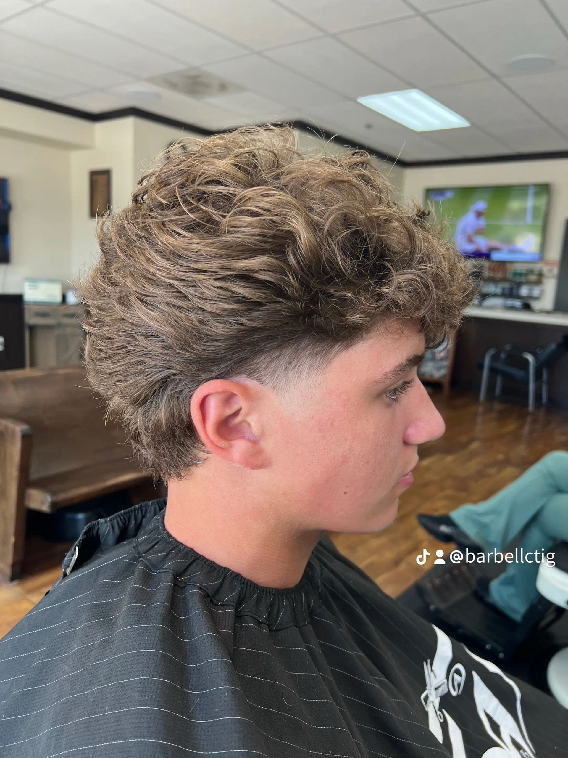Man with textured, curly brown hair, fresh haircut, skin fade on sideburns. Sitting in a barber shop.
