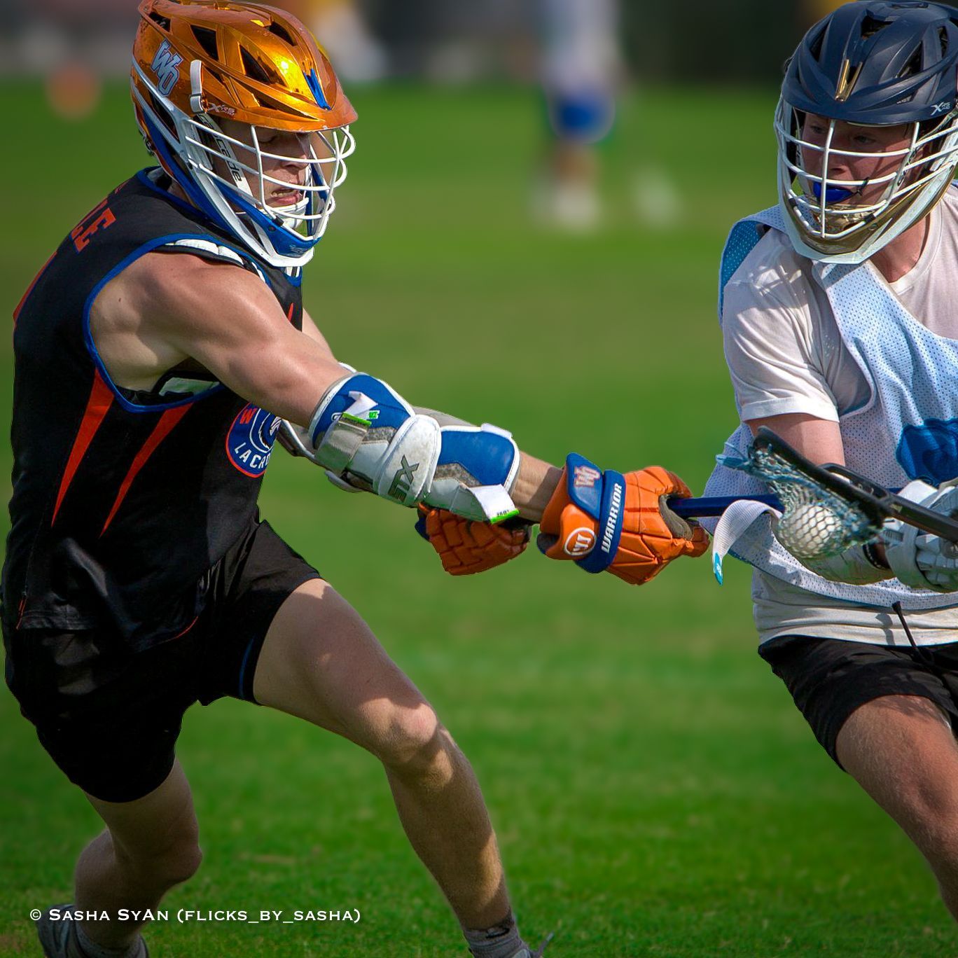 Two lacrosse players in helmets and pads tussle for the ball on a grassy field.