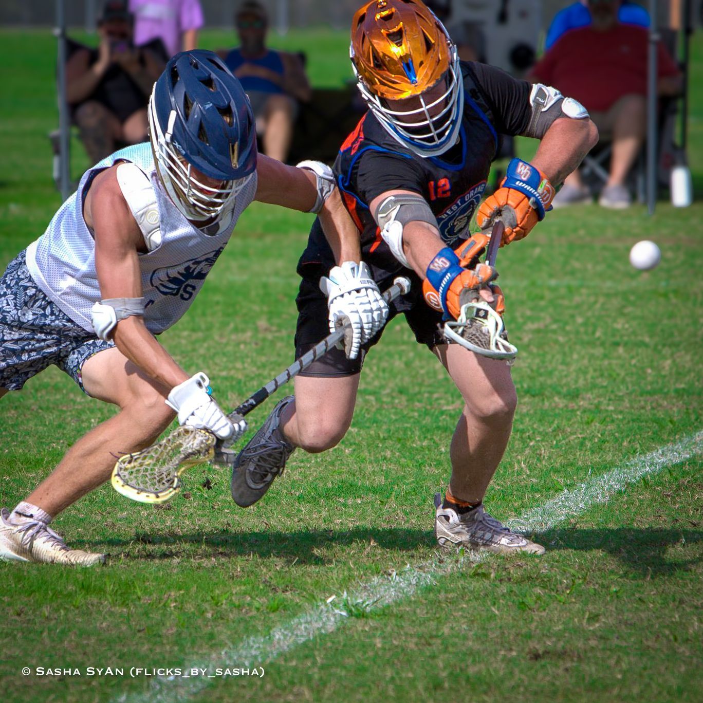 Two lacrosse players in helmets race for the ball on a grass field.