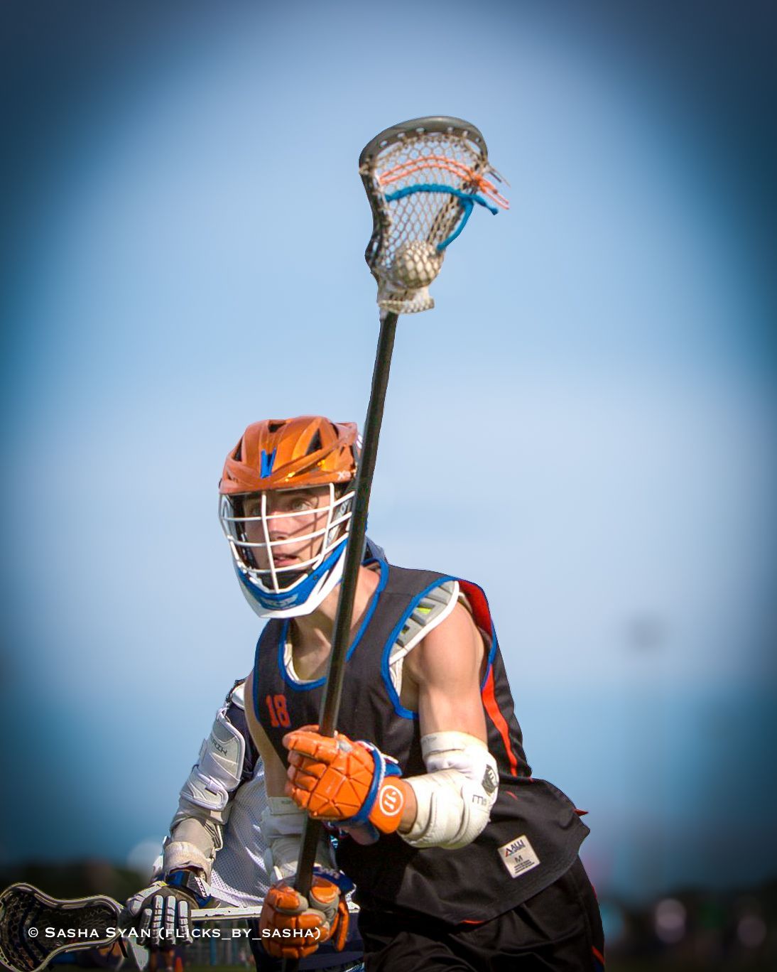 Lacrosse player in orange helmet and blue jersey, holding a stick against a clear blue sky