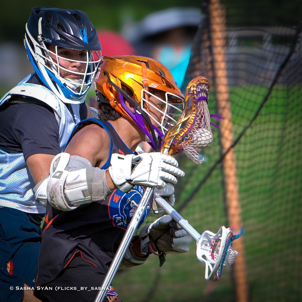 Two lacrosse players in protective gear chase the ball near the goal net outdoors.