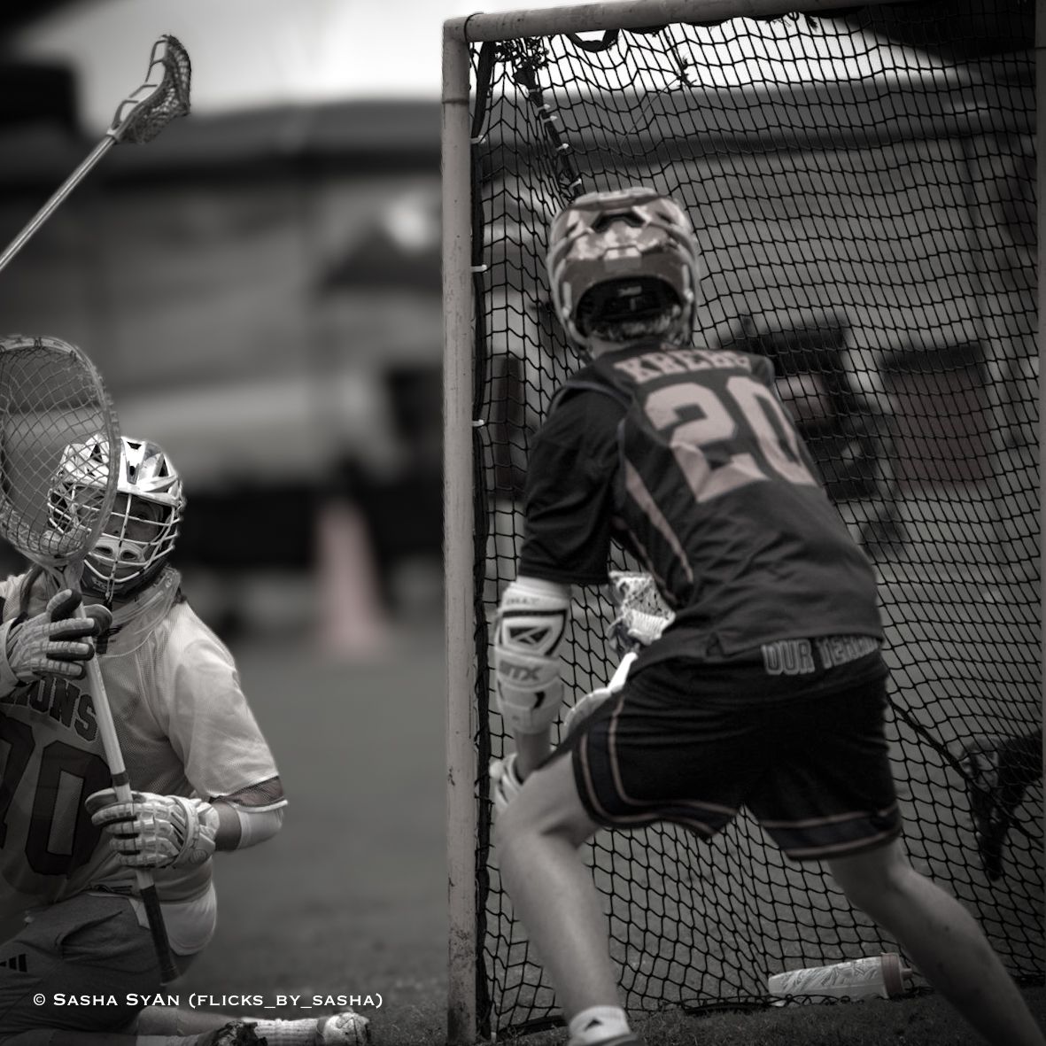 Youth lacrosse players near the goal, one in dark jersey defending against an attacker in white.