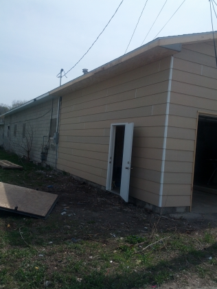 Tan building with an open white door propped against the exterior wall. Garage door visible on the right.