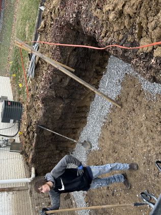 Person working in a trench, gravel base, electrical box, and wood supports present.