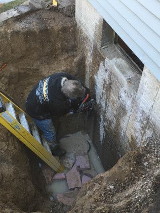 Person working in a deep trench next to a house foundation, using a tool.