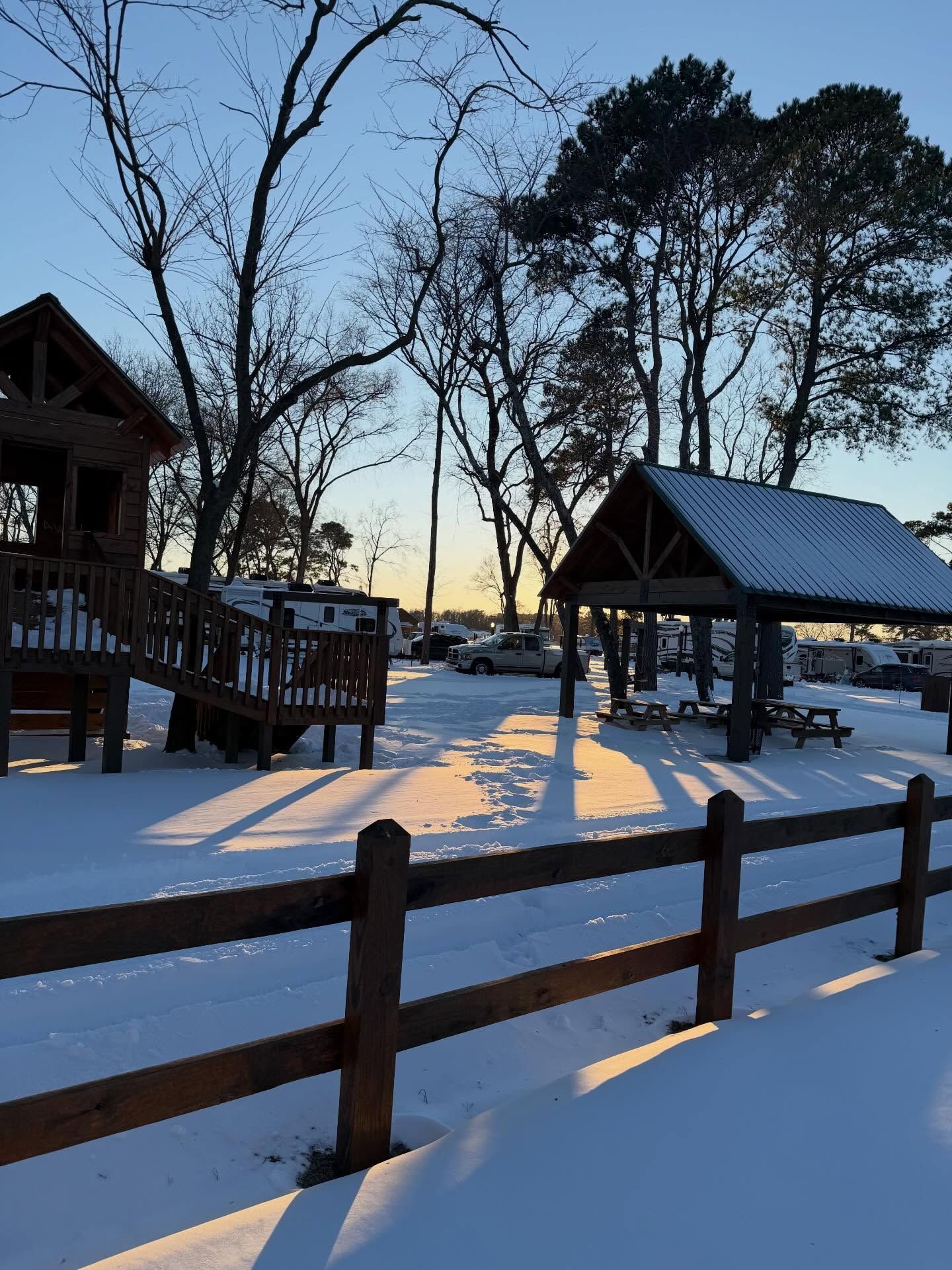 Snowy cabin yard at dusk with wooden fence, gazebo, and tall trees silhouetted against the sky