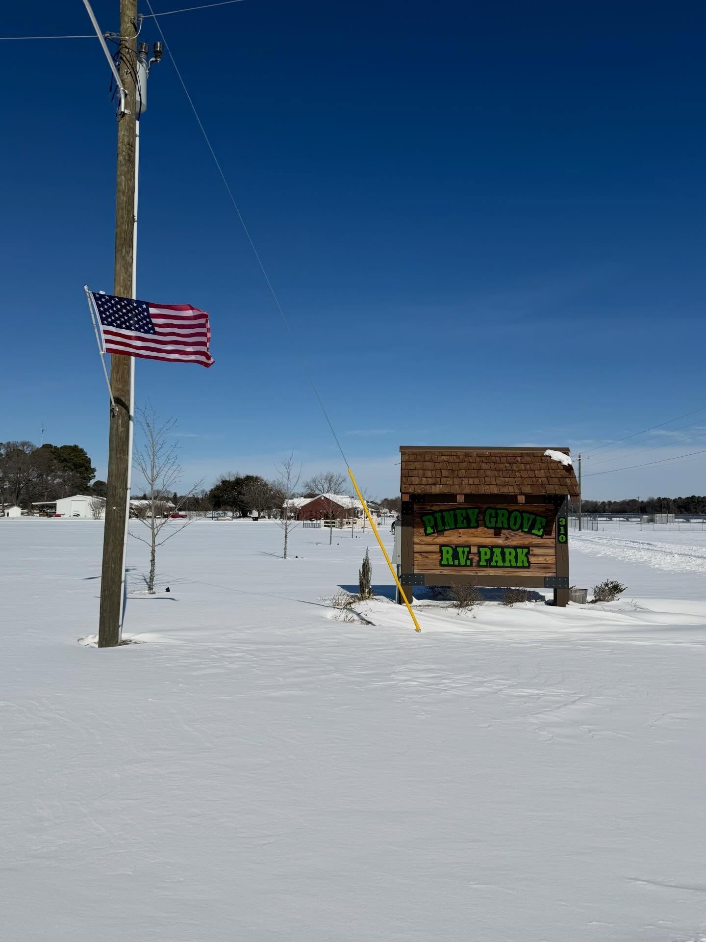 Snowy roadside scene with a U.S. flag on a pole and a small wooden sign under a clear blue sky