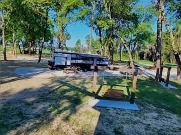 Park with trees and a long black trailer parked on a dirt road near picnic tables and a small sign.
