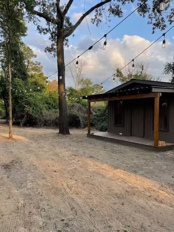Dirt path beside a small wooden cabin under string lights, with trees and a cloudy sky.