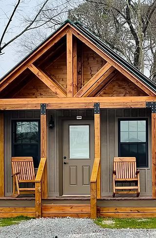 Small rustic cabin porch with a tan door, dark windows, and two wooden chairs.