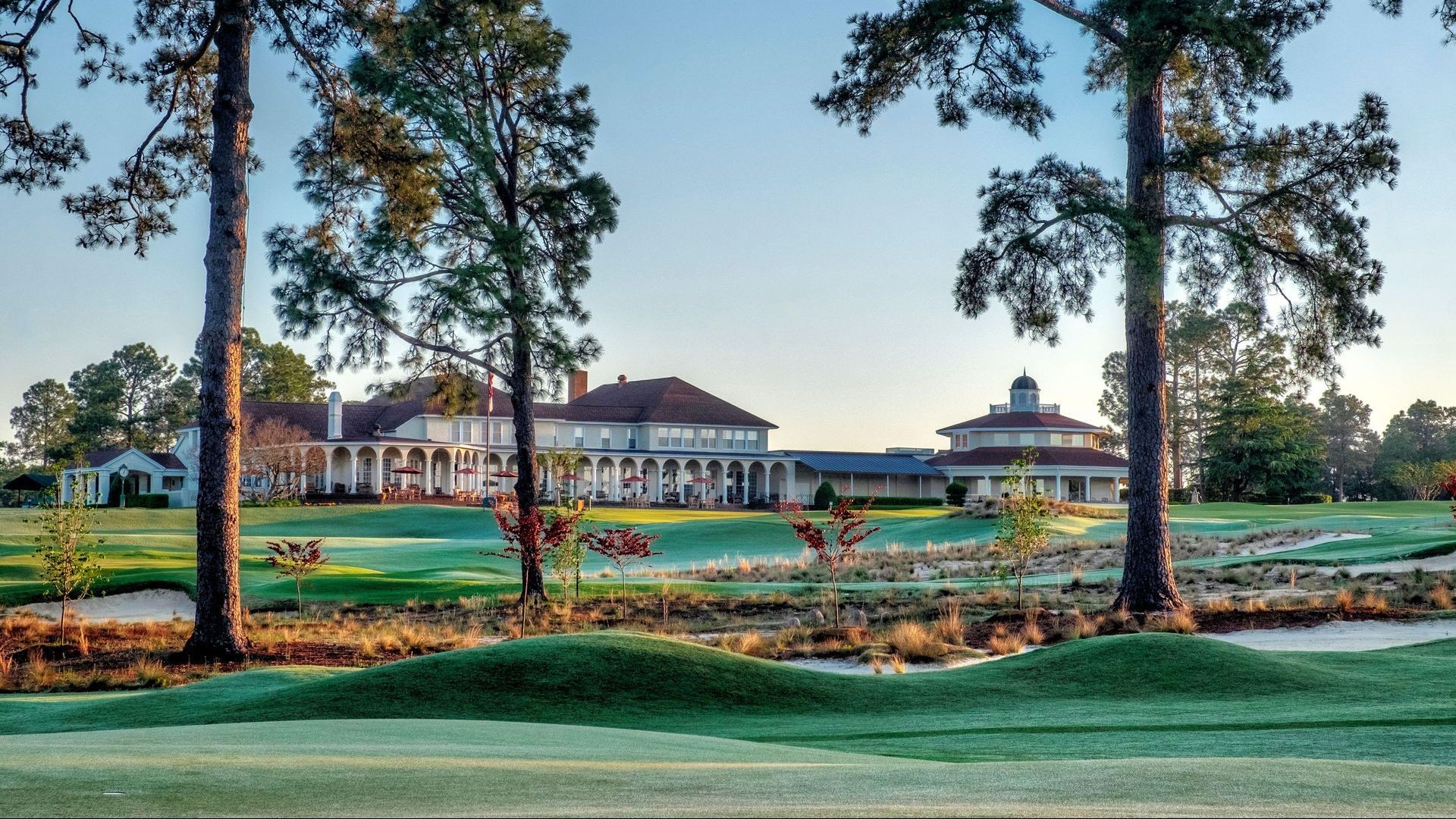 Golf course with a large, multi-story building in the background, framed by tall trees.