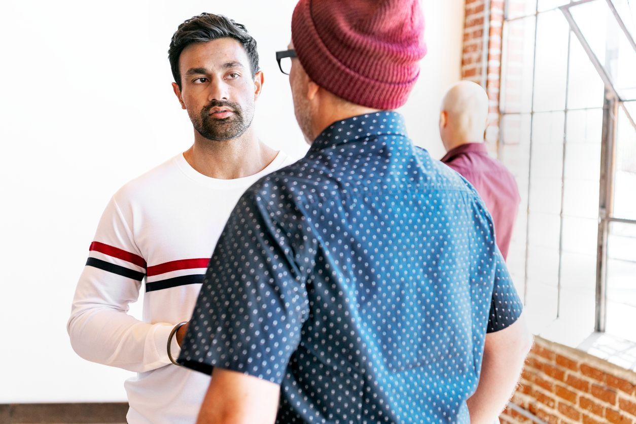 Men in conversation, standing near a window. One wears a beanie and patterned shirt, another a striped shirt.