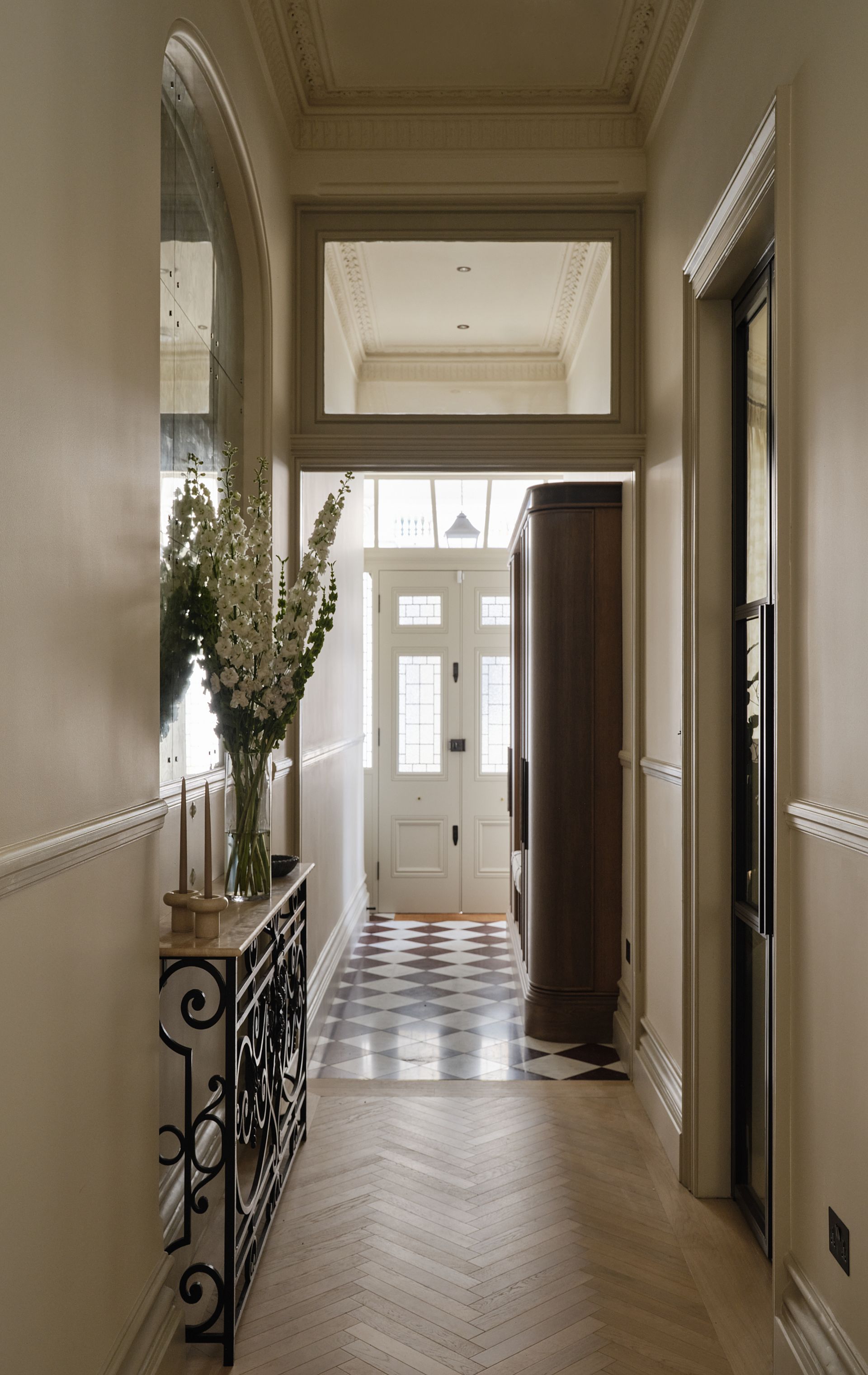Narrow hallway with herringbone wood floor, ornate mirror, and front door at the end.