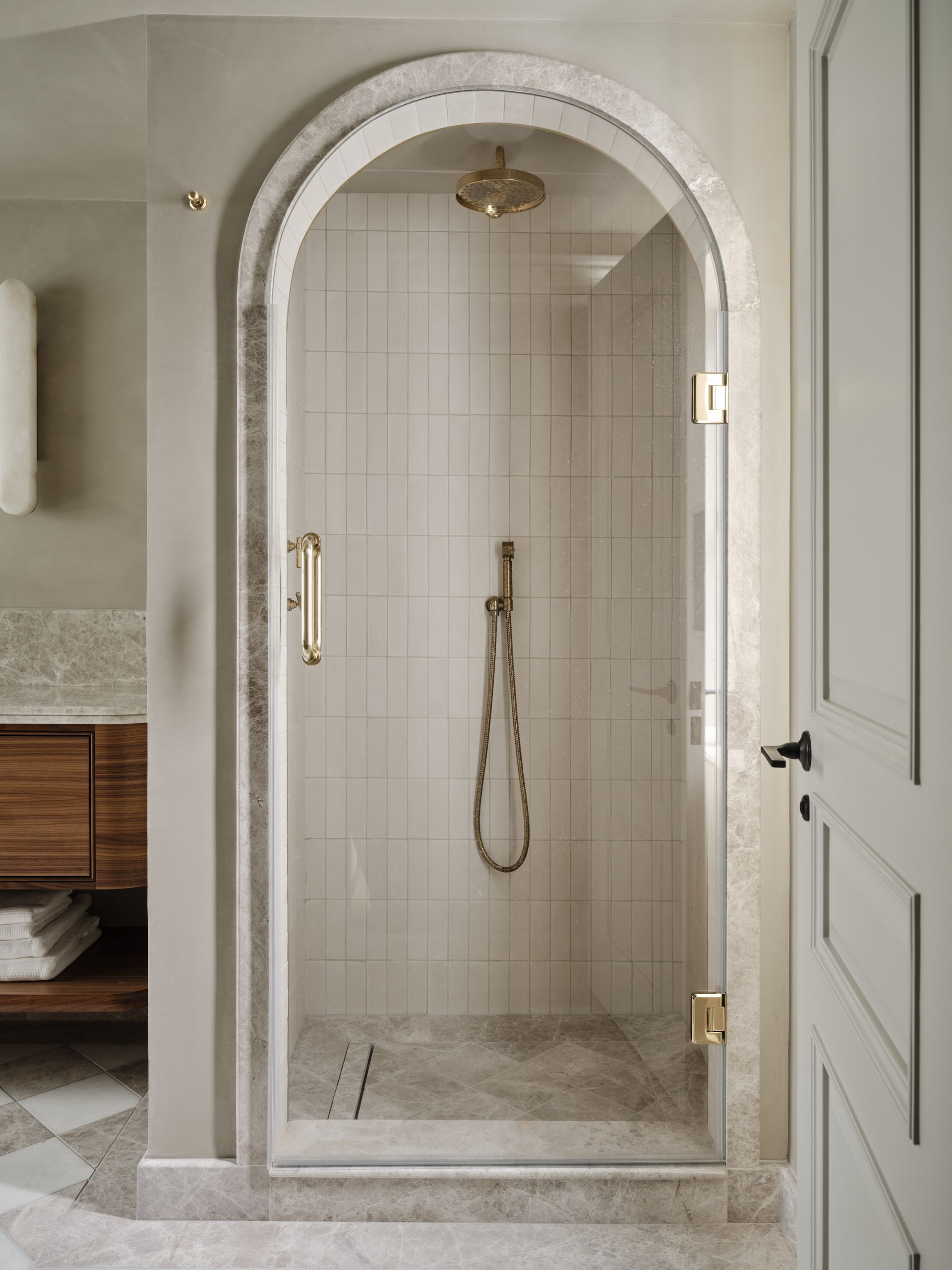 Arched glass shower enclosure in a beige tiled bathroom with brass hardware.