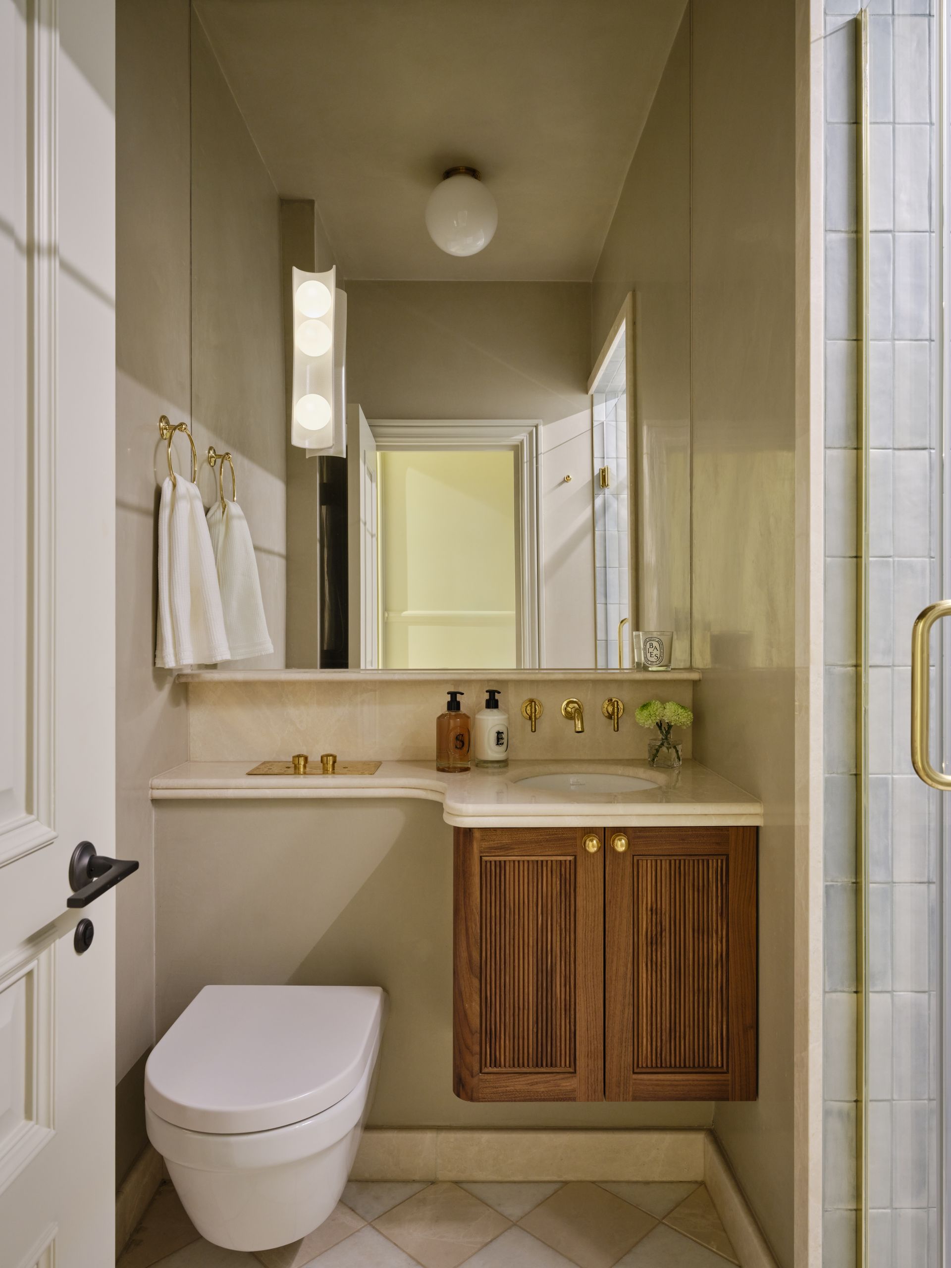 Small beige bathroom with toilet, sink, mirror, shower, and wood-front cabinet.