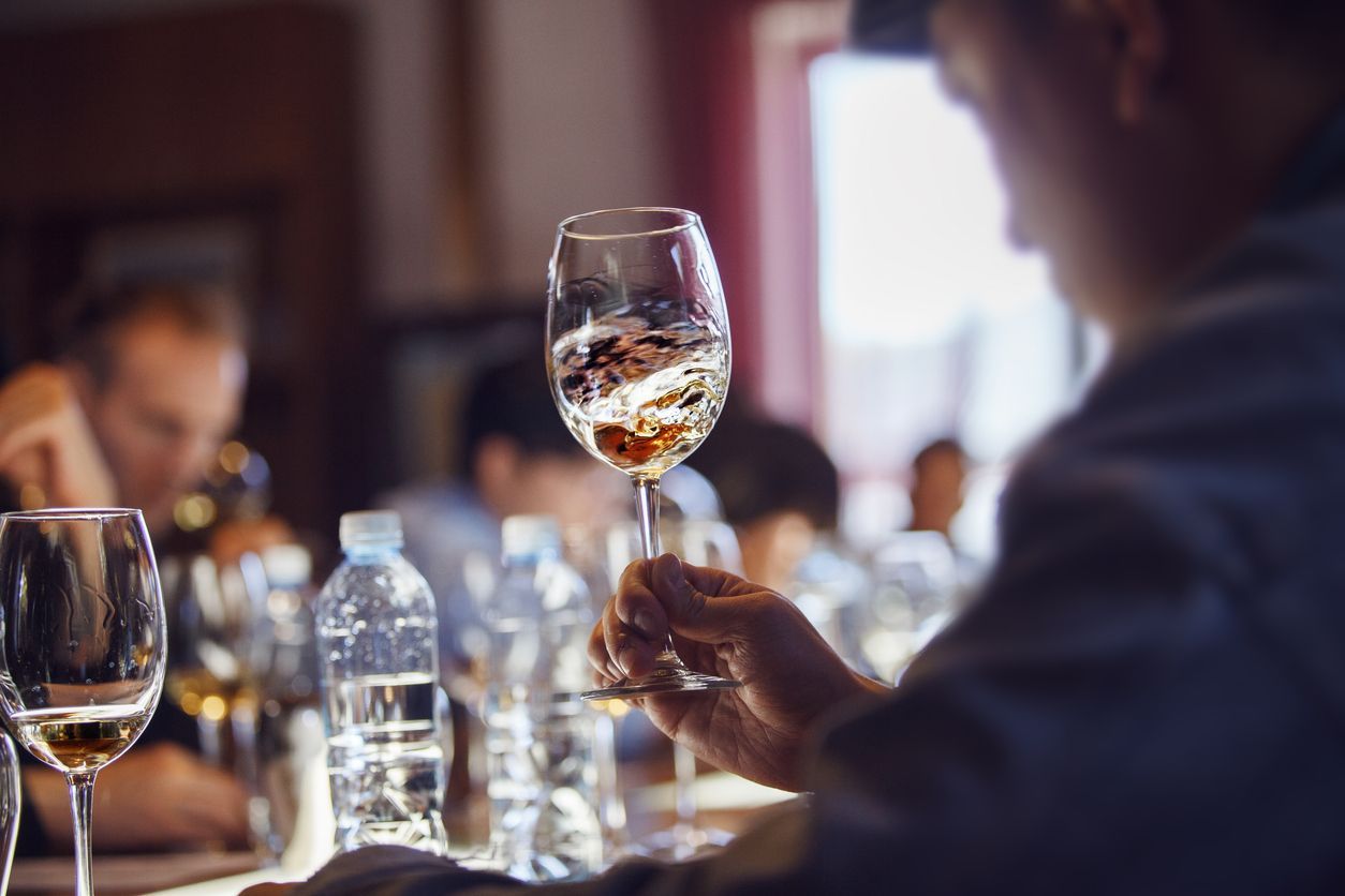 Person examining a glass of wine at a tasting event, other people, glasses, and water bottles in view.