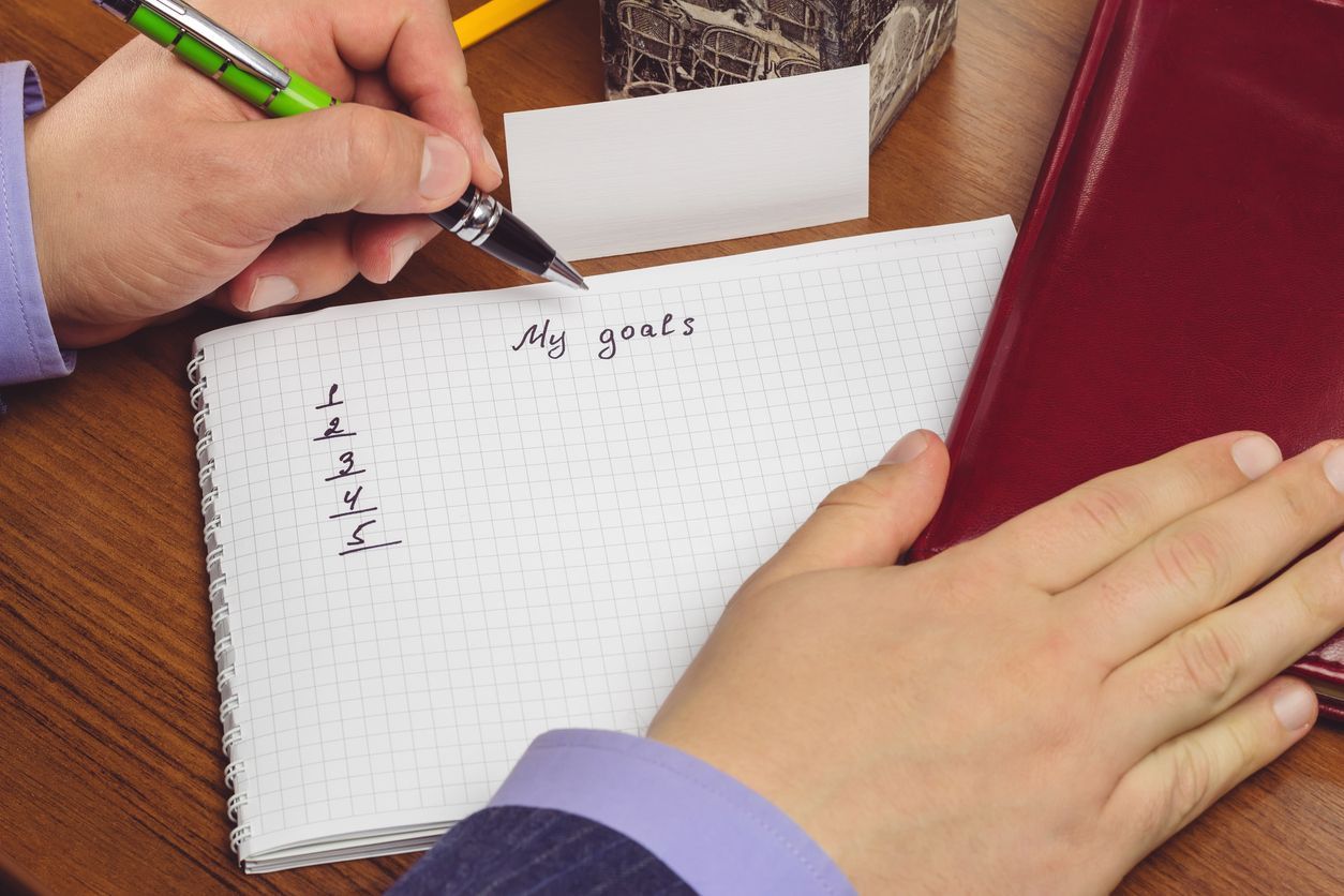 Person writing goals on a notepad with a pen. Includes a red notebook and business card on a wooden desk.