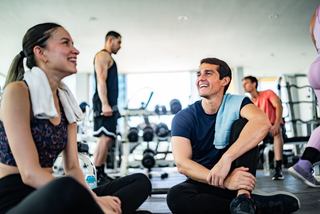 People laughing while resting on the floor in a gym. A man lifts weights in the background.