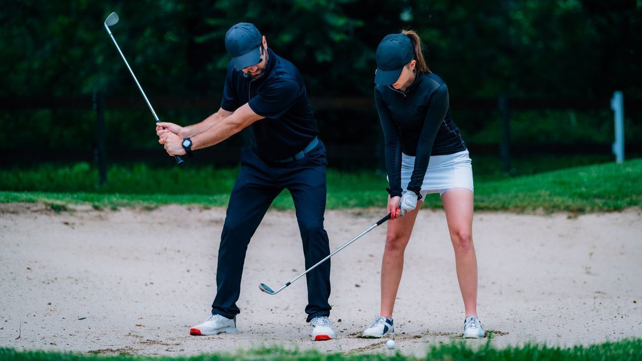 Man and woman practicing golf swing in a sand trap; both wearing dark clothing, hats.