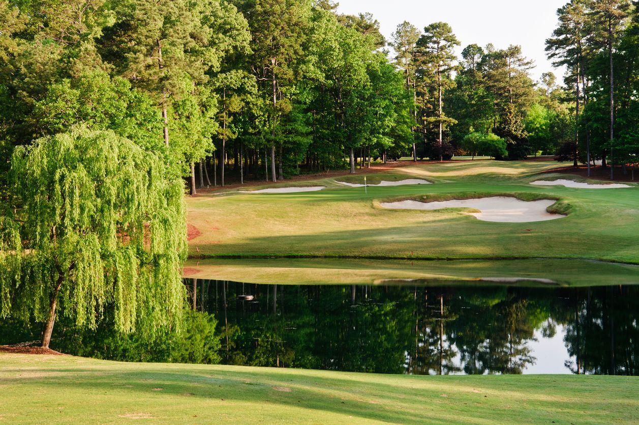 Green golf course scene with water feature, trees, and sand traps.